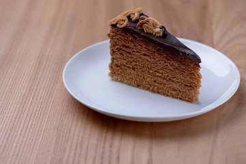 Delicious nut cake glazed with dark chocolate icing served on a white plate on light wooden table background.