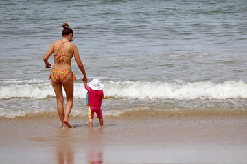 Young mom leading kid in a swimming vest to the sea. Slim woman in bikini with little child on a sandy beach, concept of single mother, family leisure, safety on a water