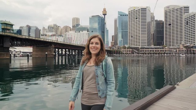 Smiling young woman walking down a pier flirtatiously looking at a camera with Sidney Australia skyline in the background