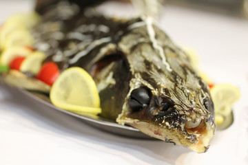 Baked sturgeon with tomatoes, lettuce and lemon slices. Black olives in its eyes. Holiday dish on the plate on white table. Stuffed seafood. Celebration event party in restaurant