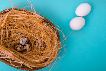 White chicken and quail eggs are in a wicker basket with straw on blue background. Easter card greeting. Flat lay. Copy space.	