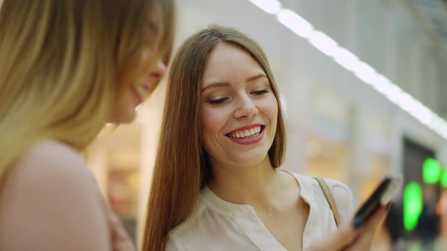 Two young and beautiful female friends with shopping bags browsing through pictures on smartphone and talking joyfully standing in shopping mall, zoom in panning shot