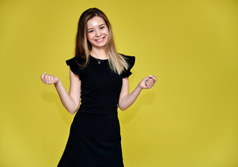 Portrait of a pretty young smiling woman on a yellow background in a black dress with long straight hair. Standing right in front of the camera, Shows emotions, talks in different poses.