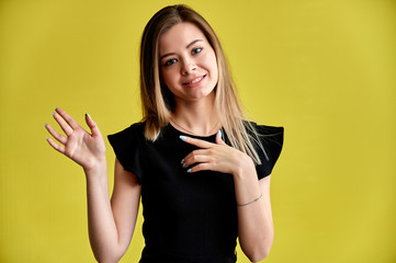 Close-up portrait of a pretty young smiling woman on a yellow background in a black dress with long straight hair. Standing right in front of the camera, Shows emotions, talks in different poses.
