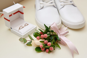 Close up of groom boutonniere with pink ribbon, white male shoes a nd wedding rings on white...