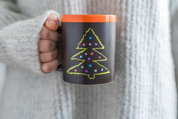 A girl in a huge knitted grey sweater holding a handmade slate black cup with painted Christmas...