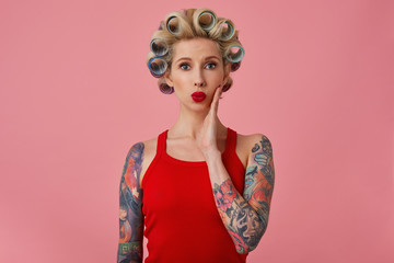 Portrait of attractive young blonde tattooed female with curlers on her head wearing evening makeup while posing over pink background, keeping hand on her face and folding her red lips