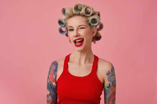 Close-up of attractive young blonde tattooed woman making hairdo while preparing for holiday celebration, winking positively at camera while standing over pink background with curlers on her hair