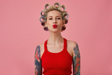 Positive lovely young tattooed female curling her blonde hair on curlers while preparing for...