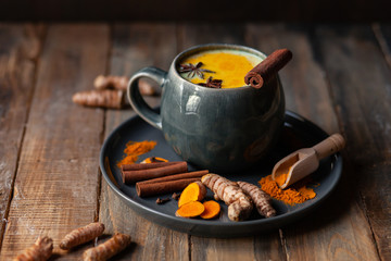 Golden milk in grey mug. Turmeric latte made with curcuma, cinnamon, anise. Healthy hot winter drink, natural, organic beverage. Close up, front view. Wooden rustic background. Raw roots as decor - obrazy, fototapety, plakaty