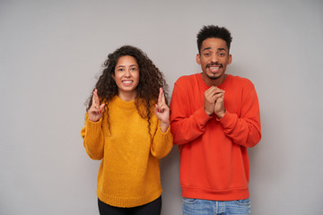 Excited pretty dark skinned young curly pair raising hopefully their hands and showing teeth while looking at camera, wearing colourful sweaters while standing over grey background