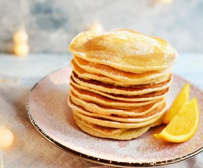 Pancakes with icing sugar and orange. Pancakes in a beautiful pink plate on a light wooden background. Delicious sweet food. Holiday pancakes and Christmas garland on the background.