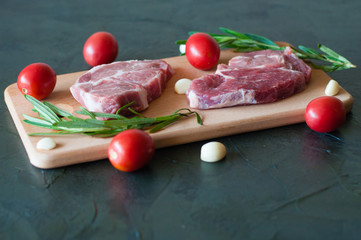 Raw pork steaks with red tomatoes, rosemary and garlic on a cutting board.