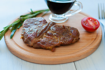 Cooked, grilled pork steak on a wooden round board, with soy sauce, rosemary and tomato, on light background with copy space