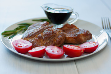 Fried, very tasty and juicy meat steaks of pork or beef with sauce, spices and tomatoes, on a gray plate, on light wooden background