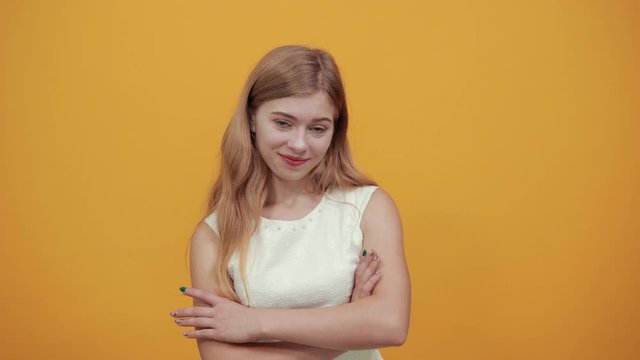 Charming blonde young woman keeping hands crossed, hugging herself, smiling, looking at camera over isolated orange background wearing white shirt. Lifestyle concept