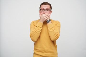 Shocked young pretty bearded male in glasses with brown short hair covering his mouth with raised palms and looking amazedly at camera, isolated over white background