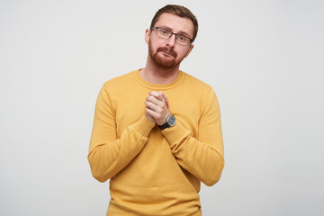 Portrait of sad young brown haired bearded guy in eyewear looking pityingly at camera with folded lips and keeping raised palms together, isolated over white background