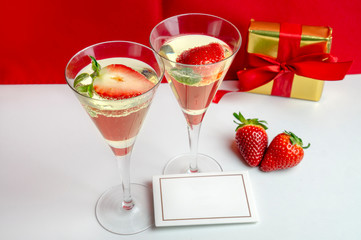 Two champagne glasses with strawberries and an empty card between them with a gift in the background . Valentine's Day concept