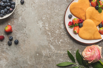 top view of heart shaped pancakes with berries on grey concrete surface near rose