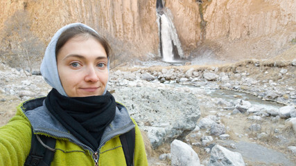 Obraz na płótnie Canvas A female traveler takes a selfie on the background of an autumn waterfall. Dressed in a warm coat, scarf and hoodie. Smiles. Travel in cold weather