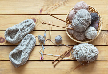 Rustic still life with handmade knitted warm slippers and accessories for knitting: balls of woolen yarn in woven basket, hooks, knitting needles on a wooden background. Flat lay, close-up, top view