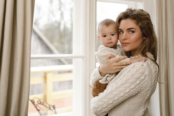 Mother and her little baby son wearing warm sweaters standing by the window