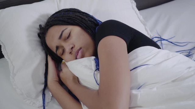 Top view of African American girl with dreadlocks sleeping and fixing blanket. Young cute teenager resting in white bed. Leisure, weekends, lifestyle.