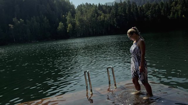 Woman lie on a sunbed in sunglasses and a boho silk shawl. Girl rest on a flood wood underwater pier. The pavement is covered with water in the lake. In the background are mountain and a forest.