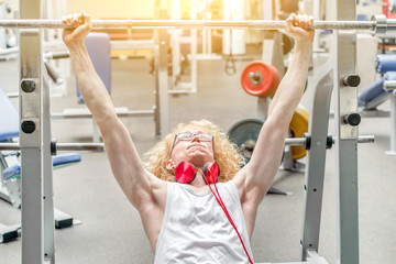 Curly blond guy in a white vest with red headphones lifting barbell with effort in the gym