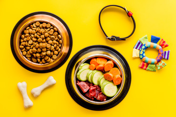 Dog feed - dry food and raw meat - near toy, collar and cheving bones on yellow background top view