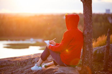 Man in hoodie writing in notebook, sitting on top of the rock. scenic view of landscape. beautiful...