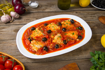 Pan seared fish with tomatoes and olives on an old rustic wooden table. Selected focus.