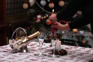 Just before the celebration: woman hand's set a candle on Christmas  decorated table with crystal glasses, wine bottle, candle and Christmas tree on the back