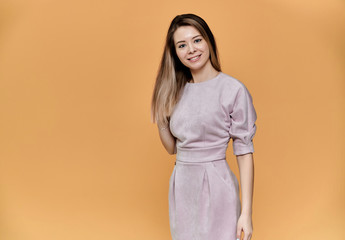 Obraz premium Portrait of a pretty young smiling woman on a beige background in a pink dress with long straight hair. Standing right in front of the camera, Shows emotions, talks in different poses.