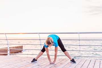 Woman with long blonde hair in blue shirt and black leggins doing stretching exercise deep side lunge withforward bend on seashore at sunrise