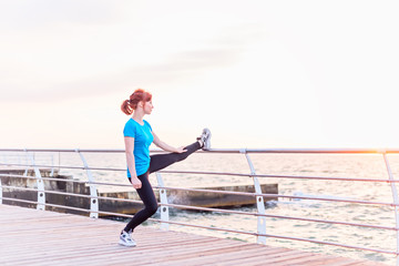 Fototapeta na wymiar Young girl with long red ponytail hair in blue shirt and black leggins doing exercise for leg stretching on handrail on seashore at sunrise