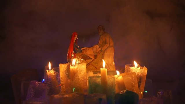 Burth of a Jesus installation inside of snow igloo house with colorful icy plafonds and burning candles in them in cold winter night. Joseph and Mary holding the newly born Jesus.