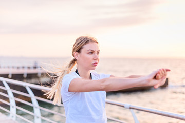 Fototapeta na wymiar Young girl with long blonde ponytail hair in white shirt doing exercise for arms on seaside at sunrise