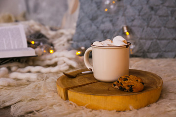 Close up shot of cup of hot cacao with bunch of marshmallow on top of wood slice tray, blurry bokeh light background. Decorative pillows, plaid blanket.