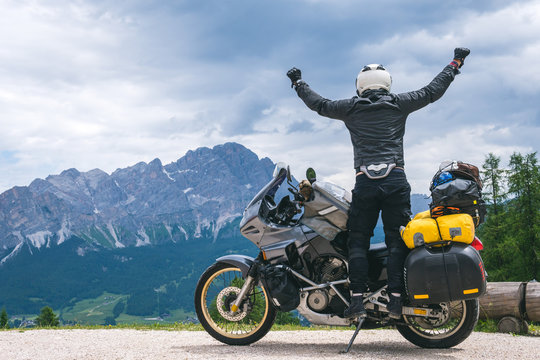 Concept of destination. conquering the top, two hands up, biker man with touring motorcycle on dirt road, "Monte Cristallo" Dolomites mountains on background, tourism travel concept, copy space, Italy
