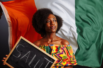 Portrait of young African woman student in national clothes holding a chalkboard with Hello word on her native language before the Ivory Coast flag