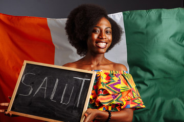 Portrait of young African woman student in national clothes holding a chalkboard with Hello word on her native language before the Ivory Coast flag
