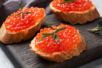 Sandwiches with red caviar on a white baguette with cottage cheese and rosemary. Dark wooden cutting Board. Grey concrete background. Close up.