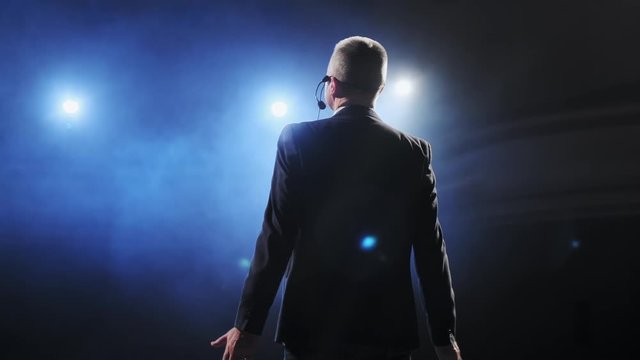 Showman. Middle Age Male entertainer, presenter or actor on stage. Arms to sides, smoke on background of spotlight. Rear view portrait of a male public speaker speaking at the microphone, pointing