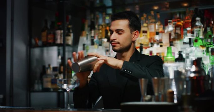 A professional bartender is preparing an alcoholic cocktail with ice cubes in a shaker to customers at the bar or disco club.