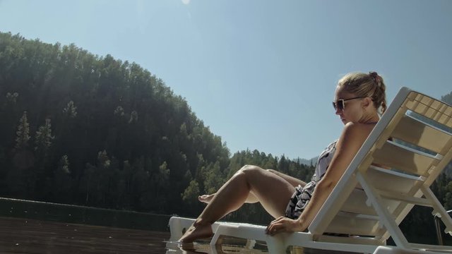 Woman lie on a sunbed in sunglasses and a boho silk shawl. Girl rest on a flood wood underwater pier. The pavement is covered with water in the lake. In the background are mountain and a forest.