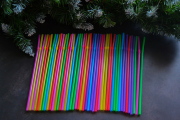 Top view of multicolored plastic cocktail straws lying in a row. In the background - green spruce branches with cones.  Festive background. Party, New year and Christmas concept. Copy space