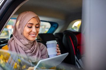 Beautiful muslim arabian woman with hijab sitting on backseat in luxury car. She drinking coffee to...