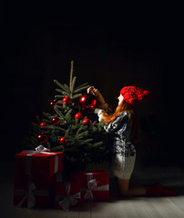 Young girl sitting celebrating decorates a christmas tree in red winter hat and presents gift boxes. New year Xmas 